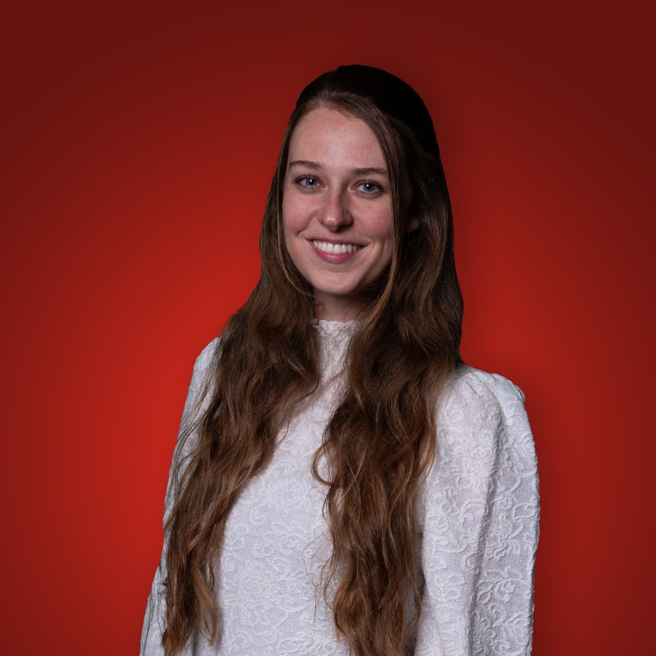 Vrouw met lang, bruin haar in een witte trui, glimlachend voor een rode achtergrond.