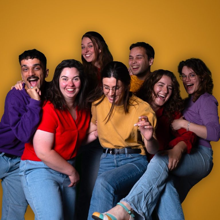 Groep van zeven mensen die vrolijk poseren voor een gele achtergrond.