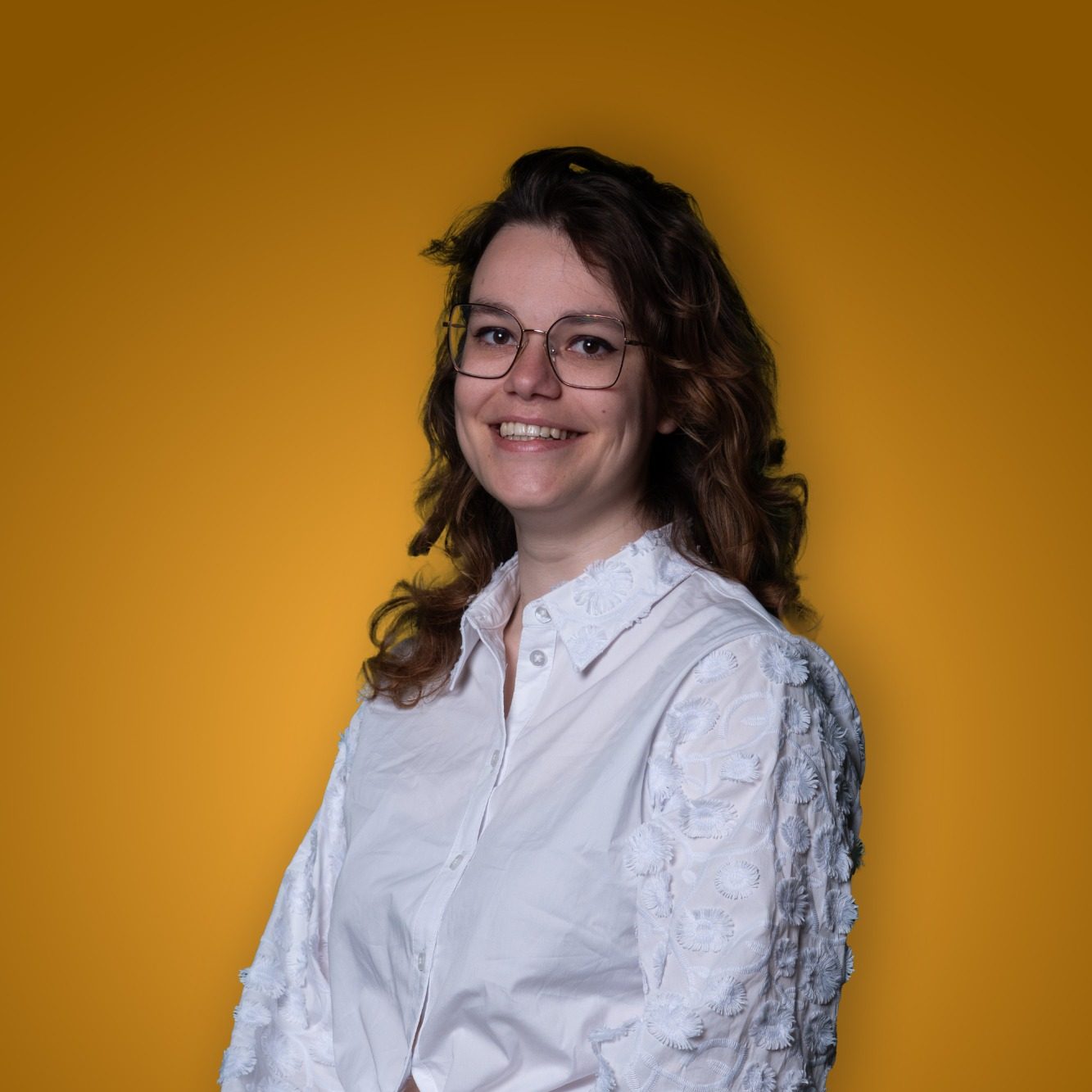 Vrouw met halflang bruin haar, bril en witte blouse, op een gele achtergrond.
