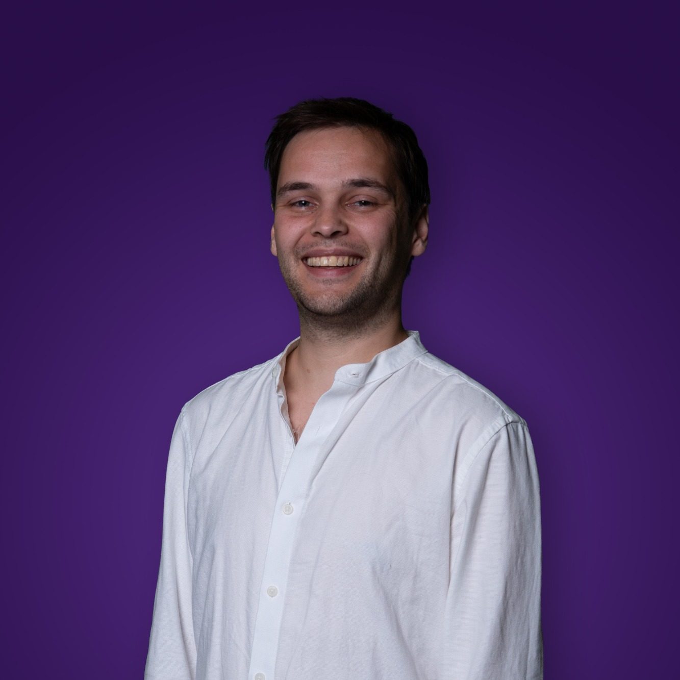 Jonge man met een glimlach, gekleed in een lichte blouse, op een paarse achtergrond.