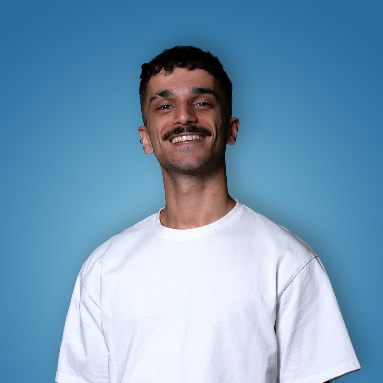 Man met een snor, glimlachend en gekleed in een wit T-shirt, tegen een blauwe achtergrond.